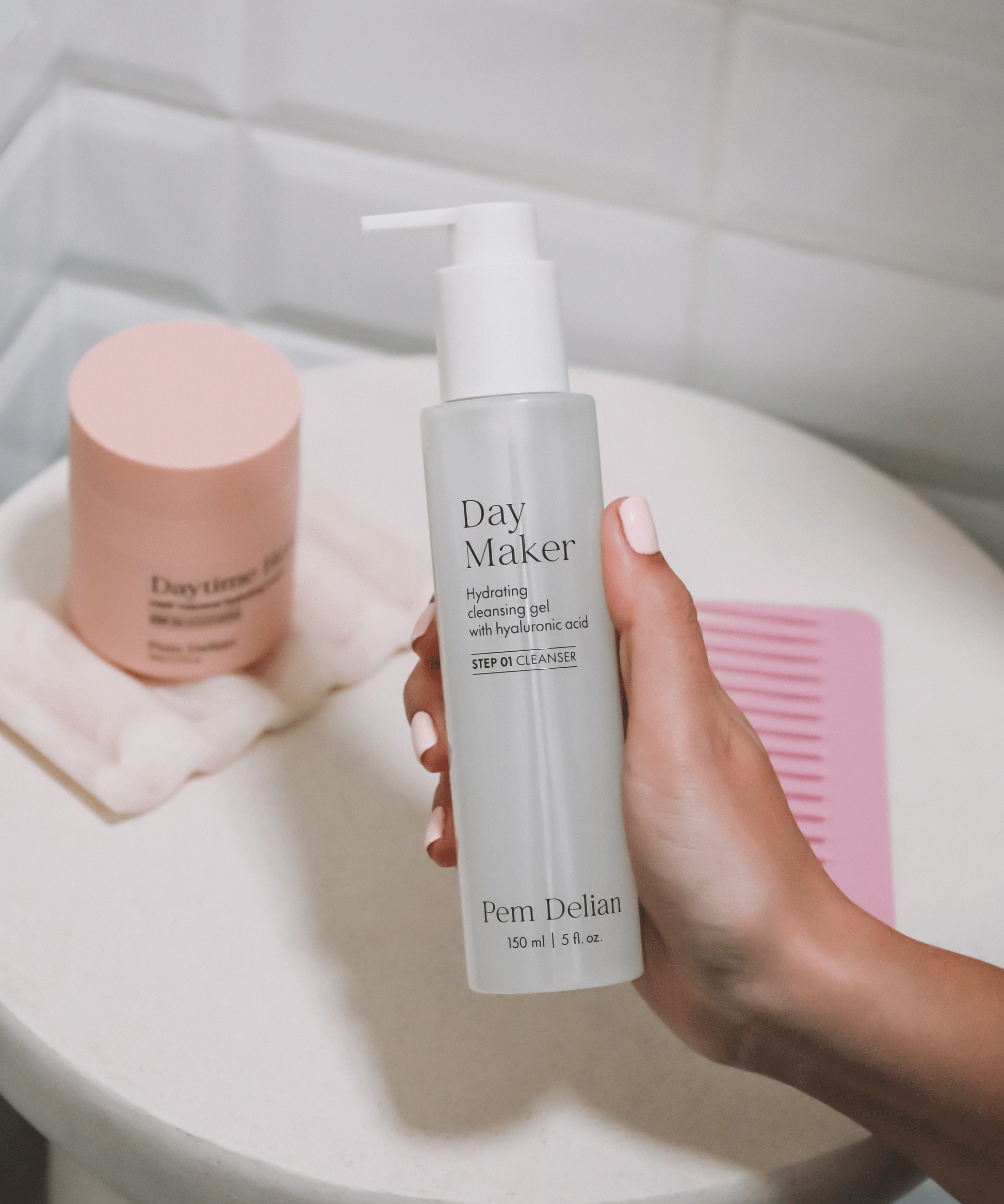 Pem Delian Day Maker Hidratáló arctisztító gél kézben tartva fürdőszobában, mögötte Daytime Hero krém és rózsaszín fésű