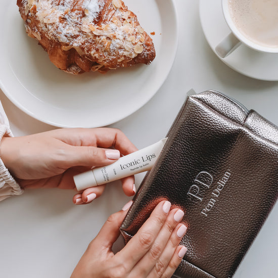 Pem Delian Iconic Lips clear ajakápolót a kézmodell az ezüst neszesszerbe tesz be, az asztalon még van egy croissant és egy bögre kávé