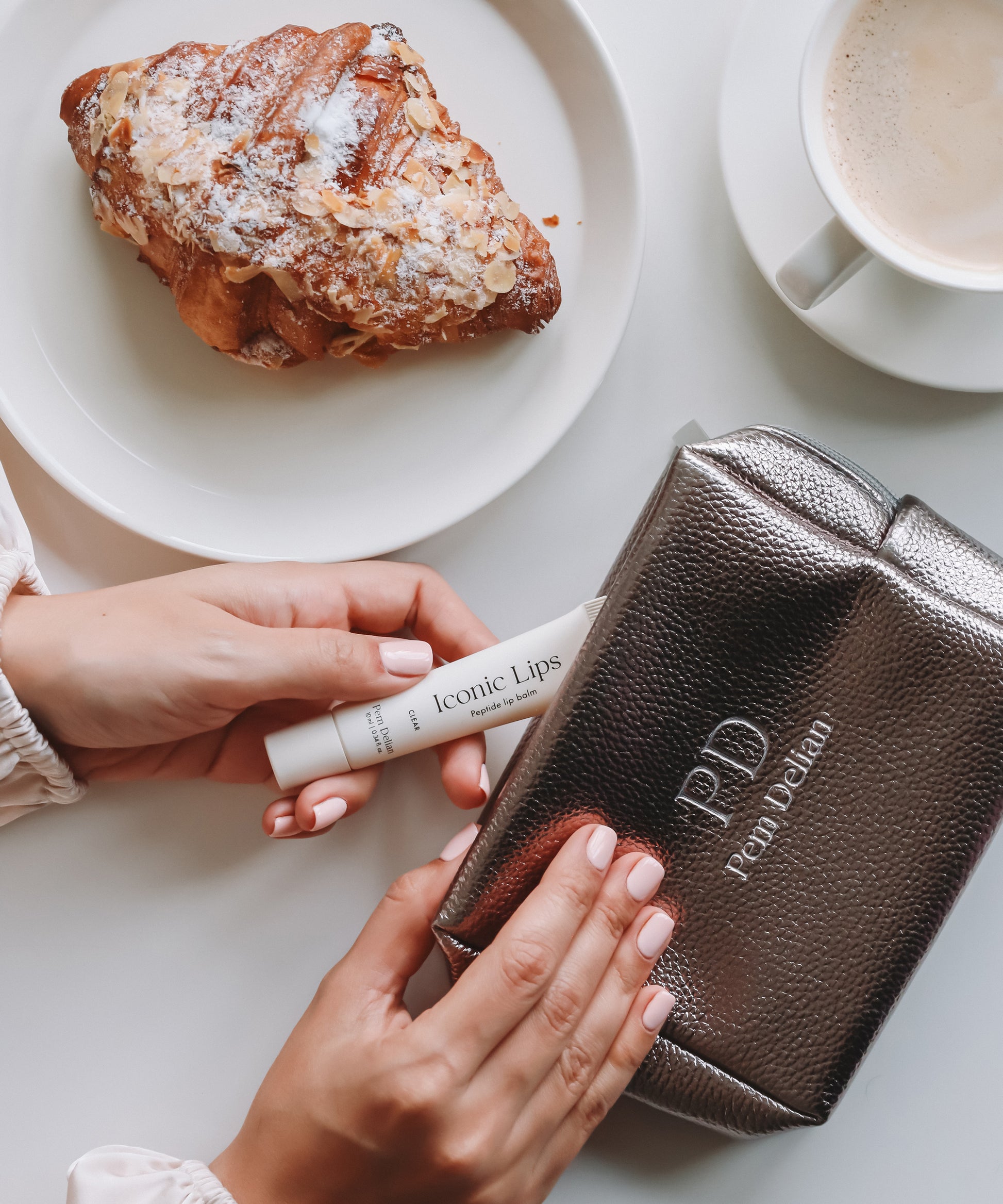 Pem Delian Iconic Lips clear ajakápolót a kézmodell az ezüst neszesszerbe tesz be, az asztalon még van egy croissant és egy bögre kávé