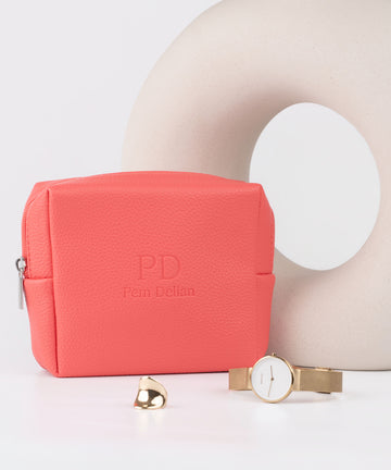 Pem Delian korall színű vegán bőr sminktáska, előtte óra és gyűrű, mögötte váza