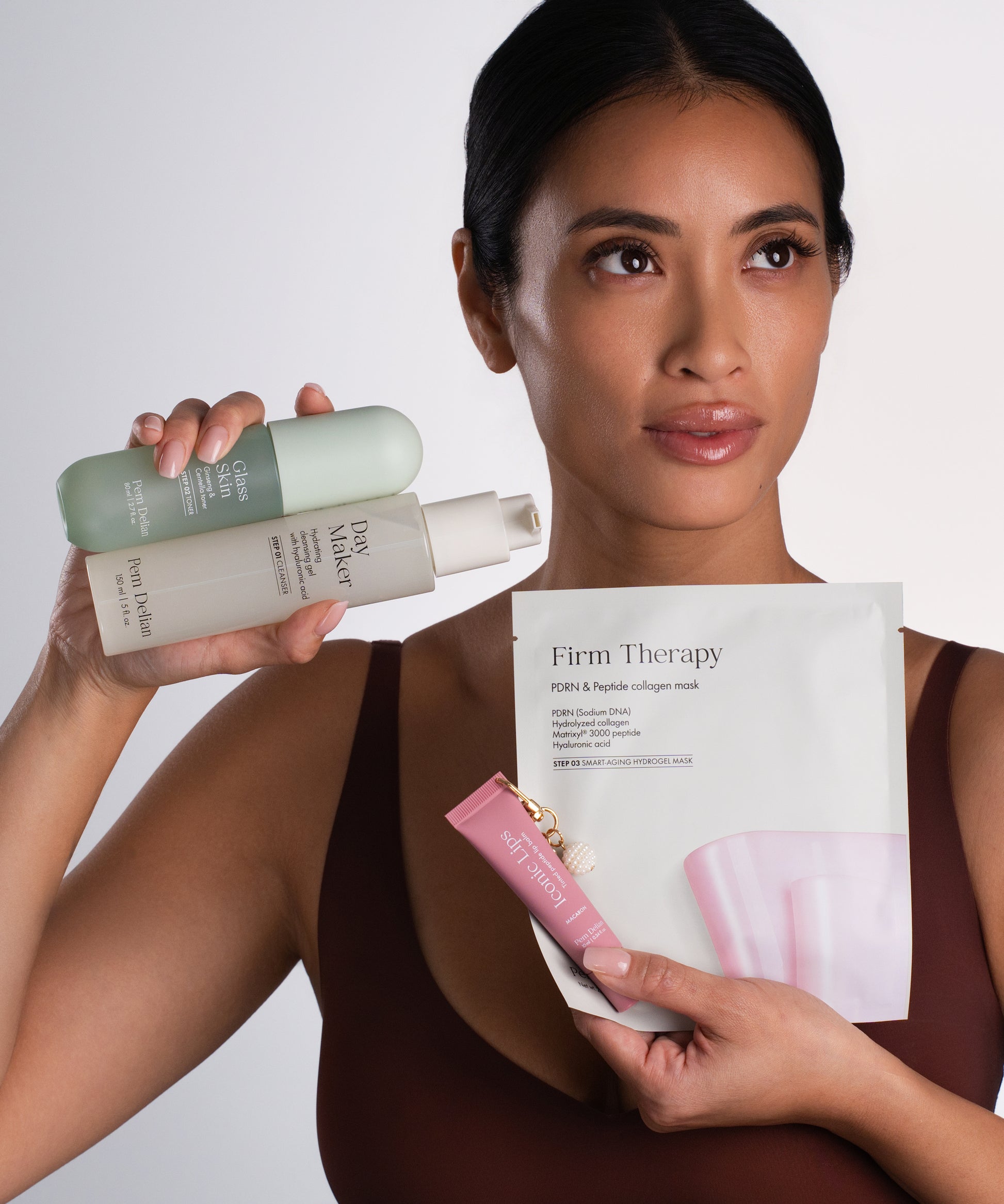 Pem Delian Glass Skin toner, Day Maker lemosó, Iconic Lips macaron ajakápoló és Firm Therapy arcmaszk a modell kezében tartva 