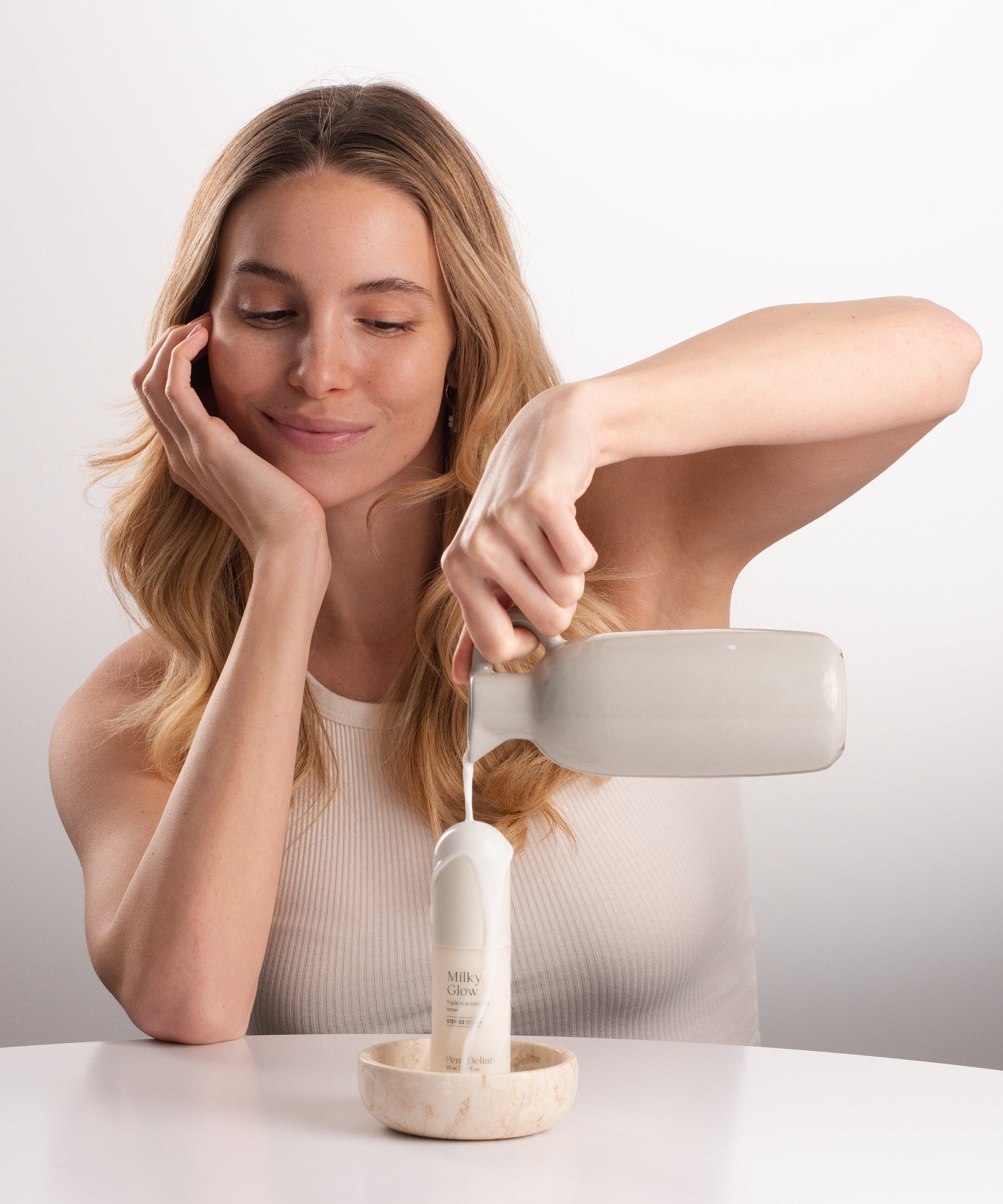 Pem Delian Milky Glow toner tejjel leöntve egy kancsóból a mosolygó modell által, fehér háttérrel