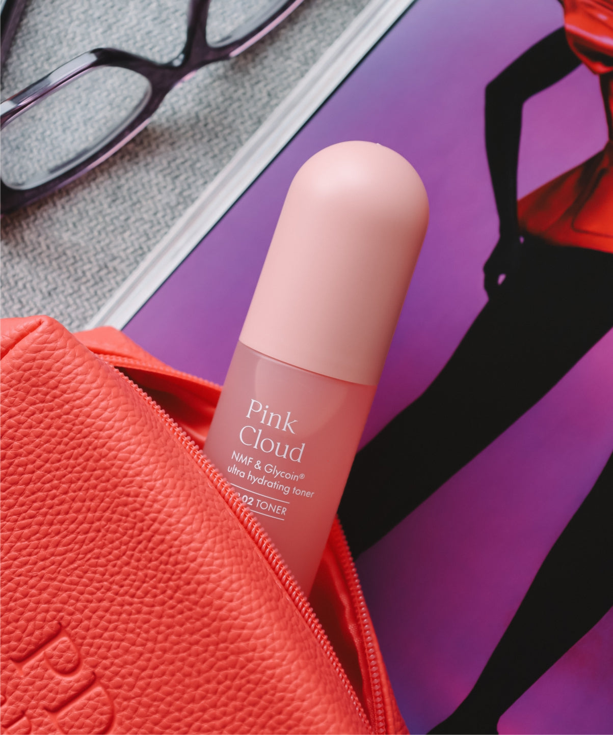 Pem Delian Pink Cloud NMF & Glycoin Ultra hidratáló toner korall színű kozmetikai táskában, mellette egy szemüveg és magazin