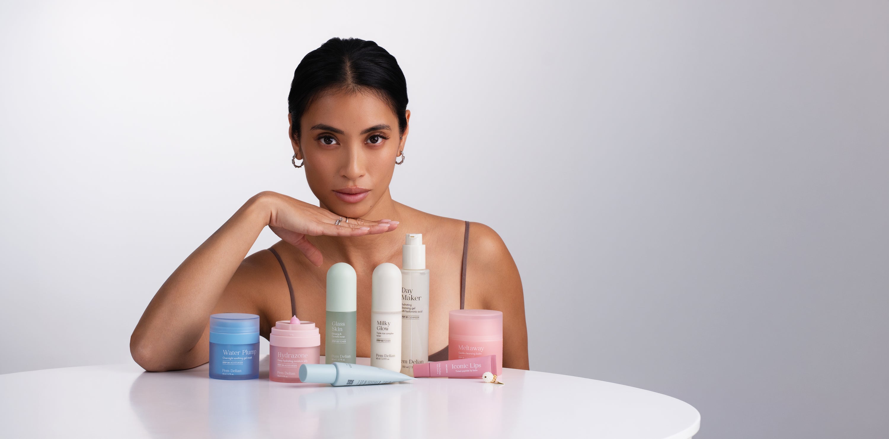 Pem Delian Water Plump, Hydrazone, Peel Power, Glass Skin, Milky Glow, Day Maker, Iconic Lips macaron és eltaway az asztalra állítva, mögötte könyököl az asztalon a sötét hajú modell